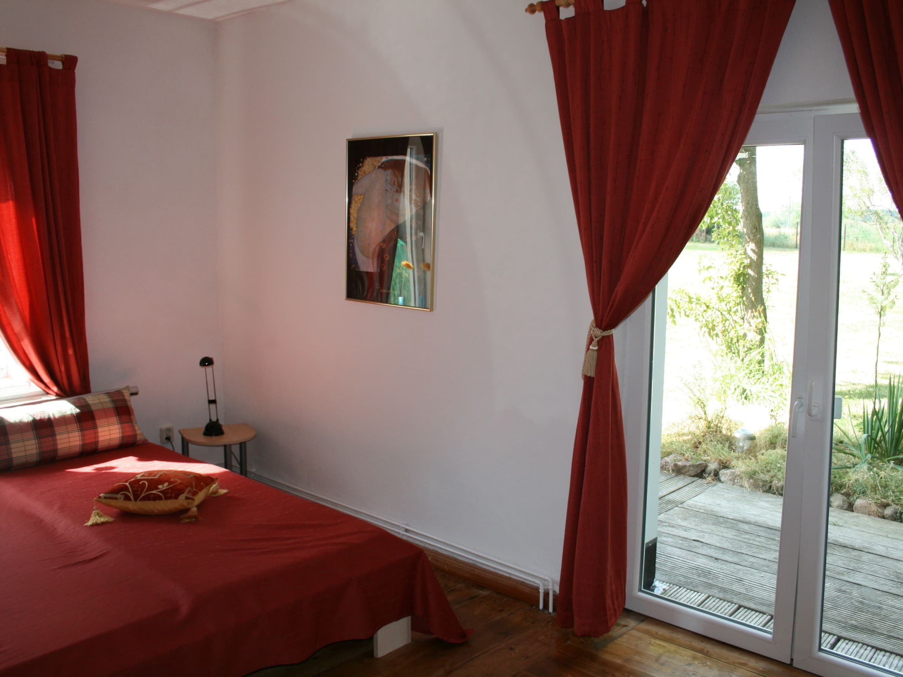 Ein Schlafzimmer mit einem Bett, einem kleinen Tisch, einer Lampe und Blick ins Freie durch eine Schiebetür.