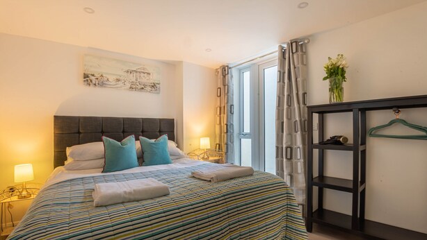A neatly made bed with striped bedding, a bedside table with a lamp, a painting on the wall, and a window with curtains.