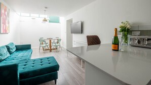 A modern kitchen with a flat-screen TV, a bar area with a bottle of champagne, and a dining area with a table and chairs.