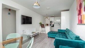 A modern living room with a teal sofa, a glass dining table, and a kitchen in the background.