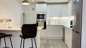 A modern kitchen with white cabinets, a built-in microwave, an oven, a sink with a scale, and a refrigerator. There are two bar stools and a pendant light above the counter.