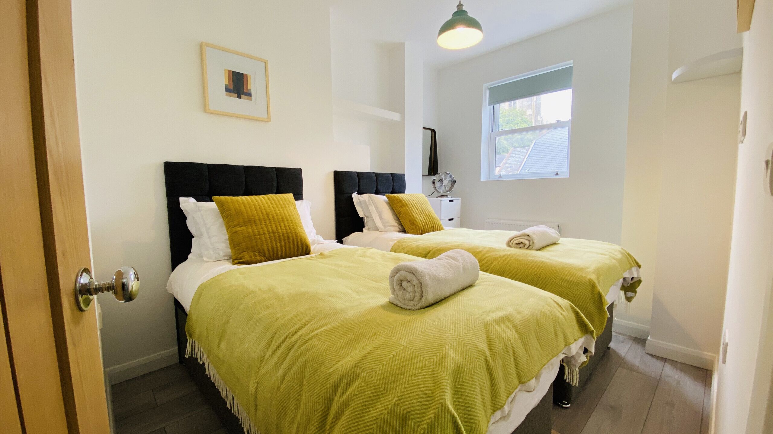 A hotel room with two beds, each with a black headboard and yellow bedding. There is a window with a view of the outside, a small picture on the wall, and a ceiling light.