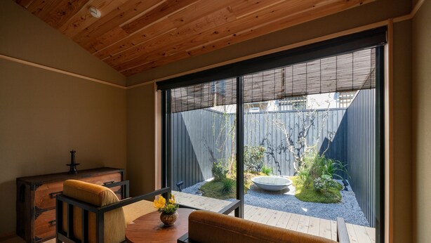 A modern interior with a wooden ceiling, a sliding glass door leading to a garden, a wooden table with chairs, and a dark wooden cabinet.