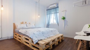 A room with a bed on a wooden pallet, a white chair, a small table, a hanging plant, and an air conditioning unit. - Boho Bangkok (Bangkok)