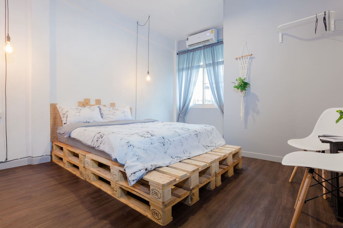 A room with a bed on a wooden pallet, a white chair, a small table, a hanging plant, and an air conditioning unit.