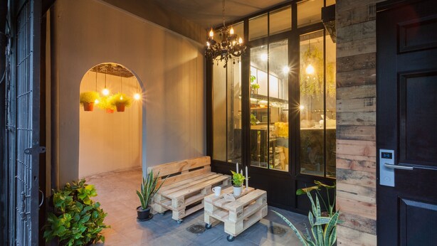 Un conjunto de banco y mesa de madera dispuesto afuera de un edificio con puerta de vidrio, plantas en macetas y un candelabro.