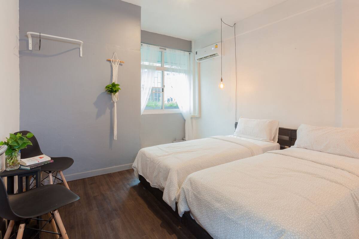 A modern bedroom with two beds, a desk with a chair, a potted plant, and a window with curtains.