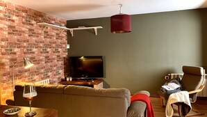 Ein Wohnzimmer mit Backsteinwand, Flachbildfernseher, Couchtisch aus Holz, einer Couch, einem Sessel und einer Pendelleuchte.