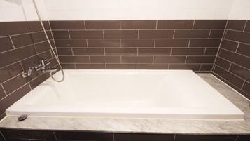 Una bañera rectangular con grifo y ducha de cromo, frente a una pared revestida de baldosas oscuras.