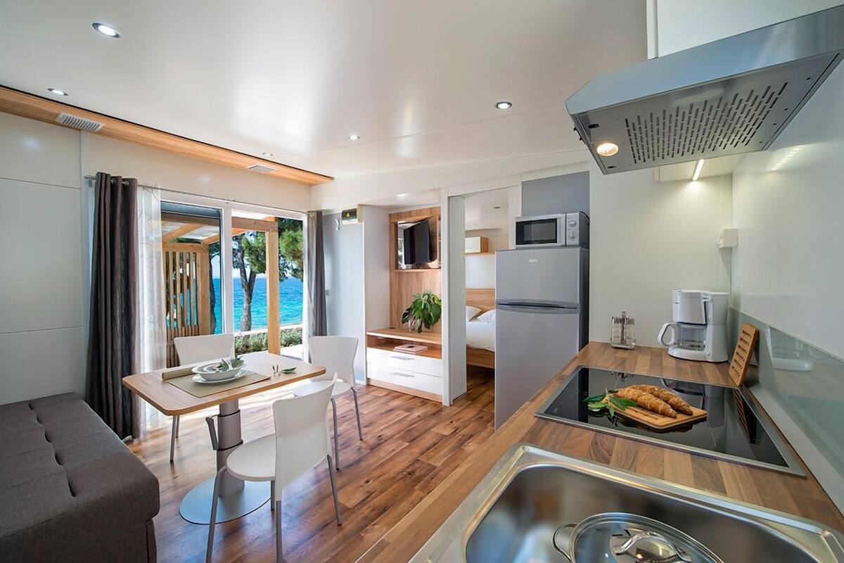 Eine moderne Küche mit integriertem Mikrowellenherd, einer Kaffeemaschine und einer Frühstückstheke. Ein Essbereich mit Tisch und Stühlen bietet einen Blick auf das Meer durch große Fenster.