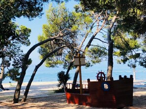 Modelo de navio pirata de madeira, com volante e boia salva-vidas, rodeado por árvores e uma praia.