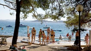 En strand med folk som solar och badar, en pool och båtar i bakgrunden.