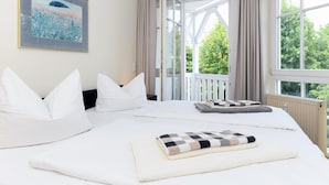 Een hotelkamer met een groot bed, wit beddengoed, uitzicht op groen door open ramen en een schilderij aan de muur.