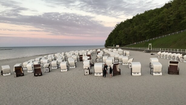 Een strand met rijen witte tenten, voorzien van nummers, een zandstrand en een bebost heuvel in de verte.
