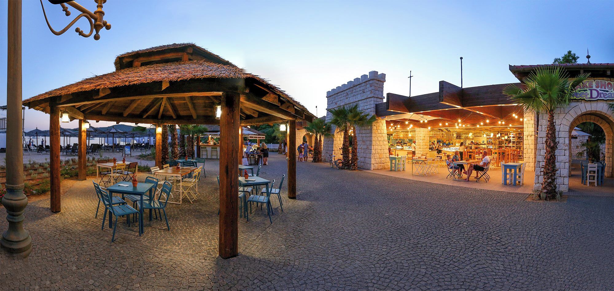 Un espace extérieur aménagé pour les repas, avec des tables et des chaises, un espace de repos couvert et un bâtiment doté d’une façade en pierre.