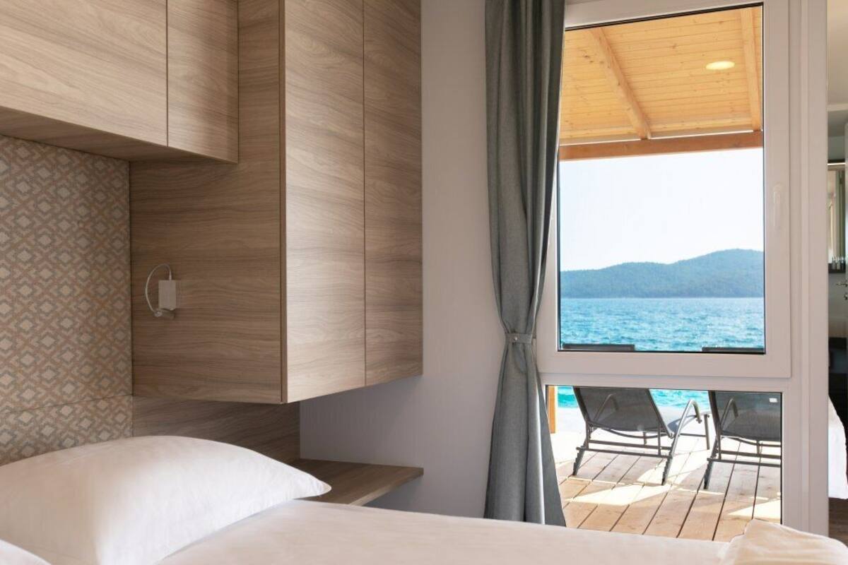 Een slaapkamer met een bed, een houten garderobe, een raam met uitzicht op zee en een balkon met stoelen.