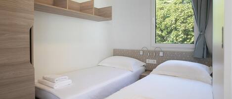 A hotel room with two single beds, a wooden wardrobe, a window with a view of greenery, and a power outlet on the wall.