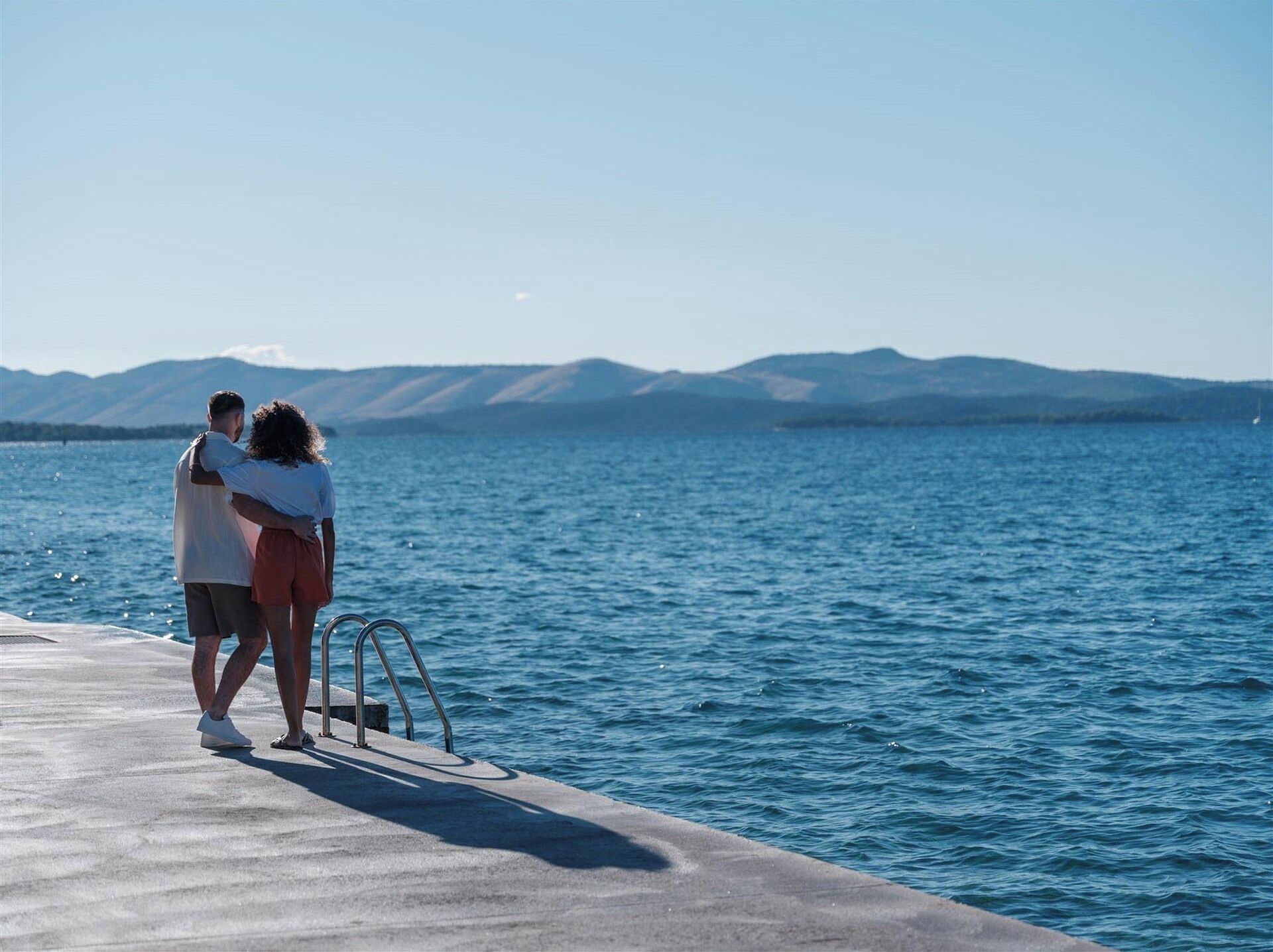 A couple embracing on a concrete pier overlooking a calm body of water with distant mountains.