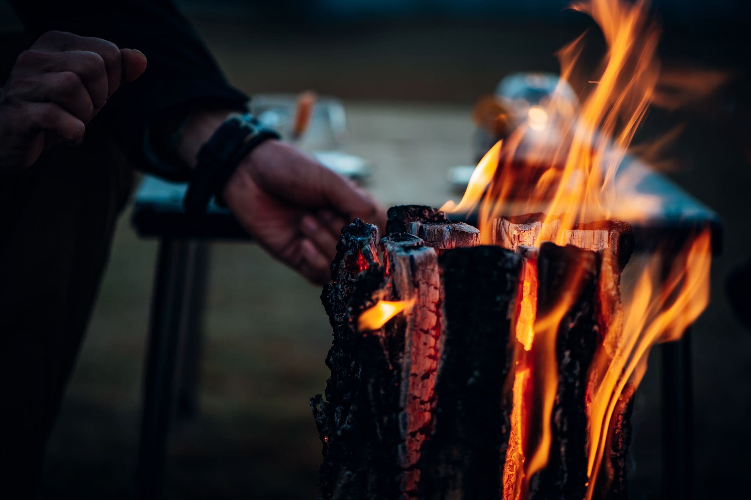 Un gros plan sur du bois en train de brûler, avec des flammes, une main et un arrière-plan flou.