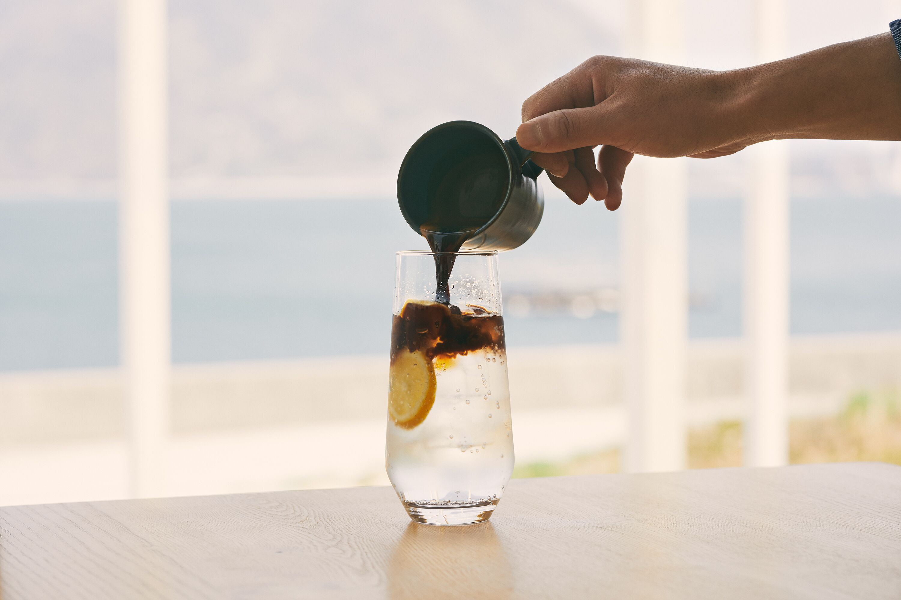 Una mano sirve un líquido oscuro en un vaso transparente con cubitos de hielo y una rodaja de limón, sobre una mesa de madera con vista al mar y montañas al fondo.