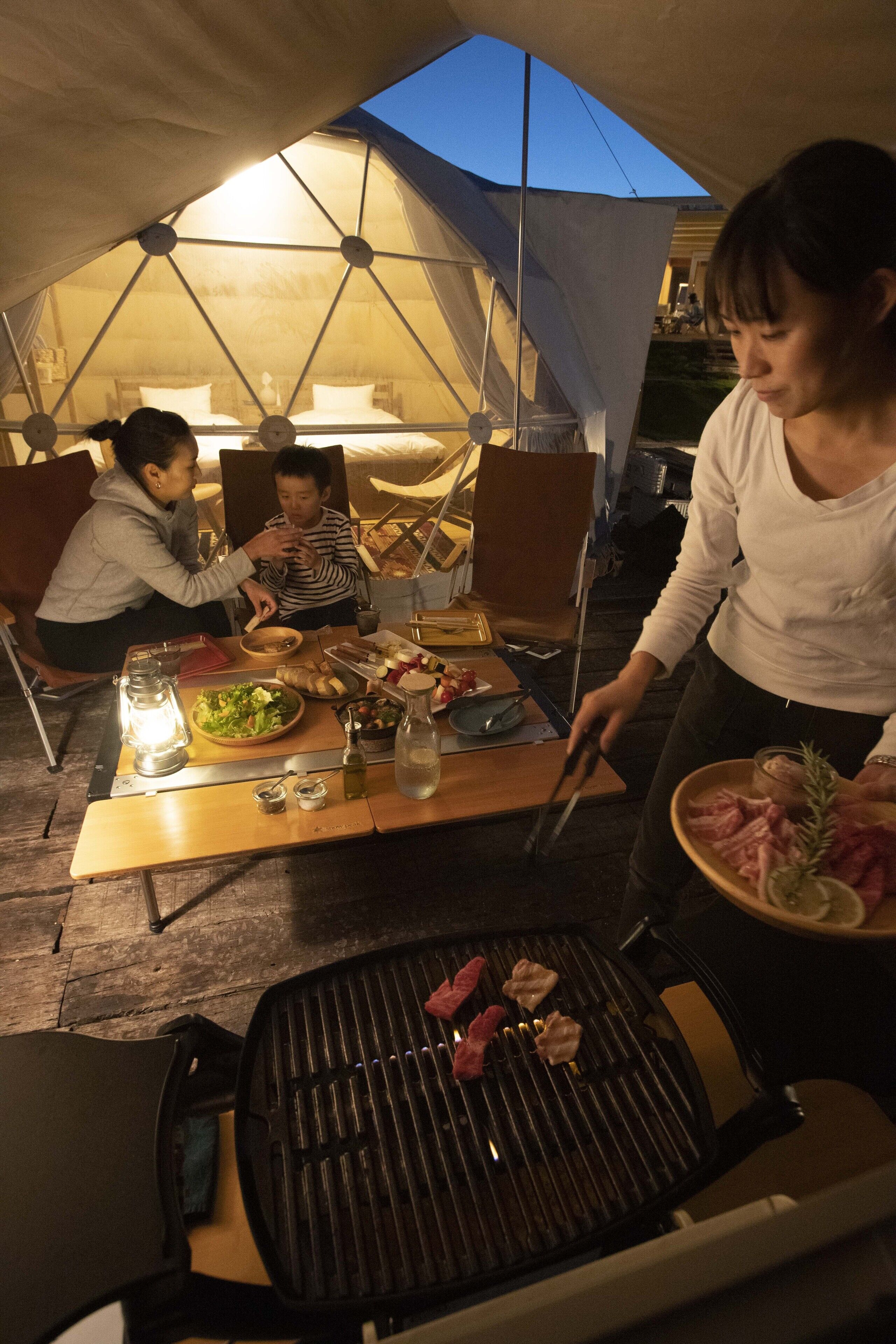 A tented outdoor dining area with a grill, table with food, and people dining.