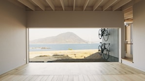 Habitación amplia con piso de madera y un gran ventanal con vista a una playa y una montaña.