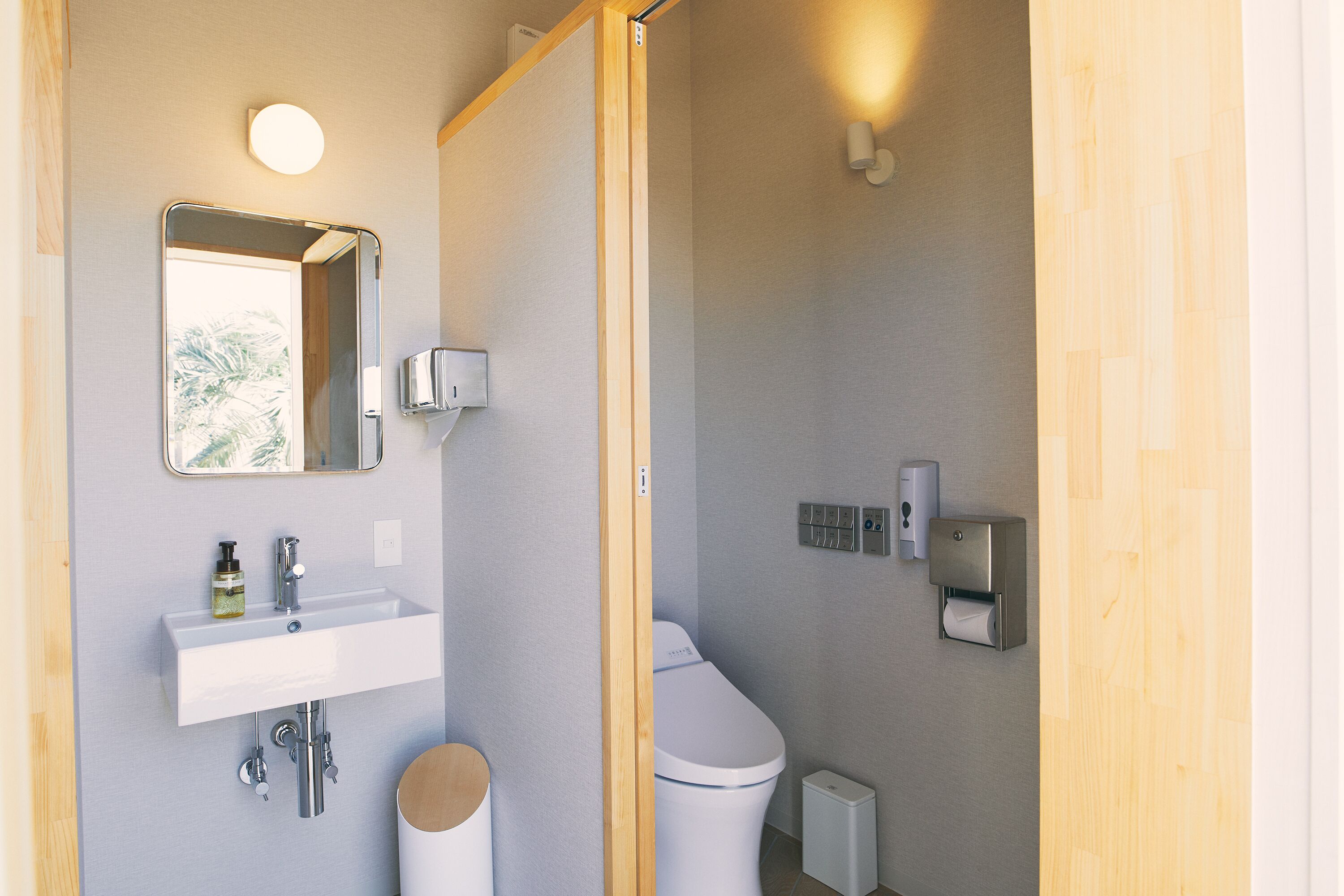 A modern bathroom with a white sink, mirror, and toilet. There are wall-mounted soap dispensers and a small trash bin.
