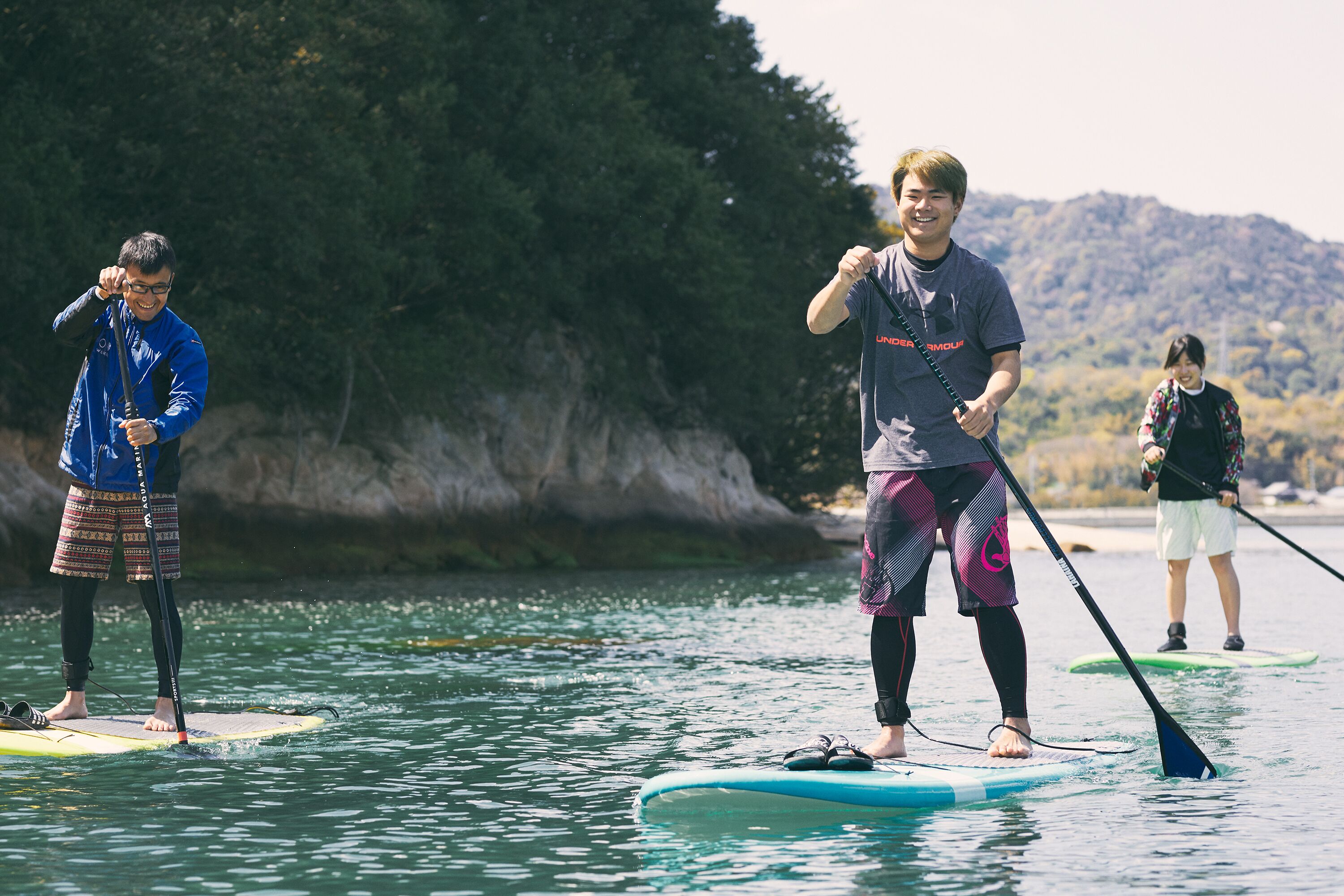 Três pessoas praticando paddleboard em águas calmas, com margem rochosa e árvores ao fundo.
