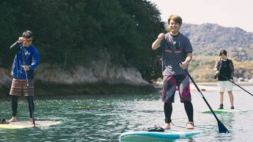 Três pessoas praticando paddleboard em águas calmas, com margem rochosa e árvores ao fundo.