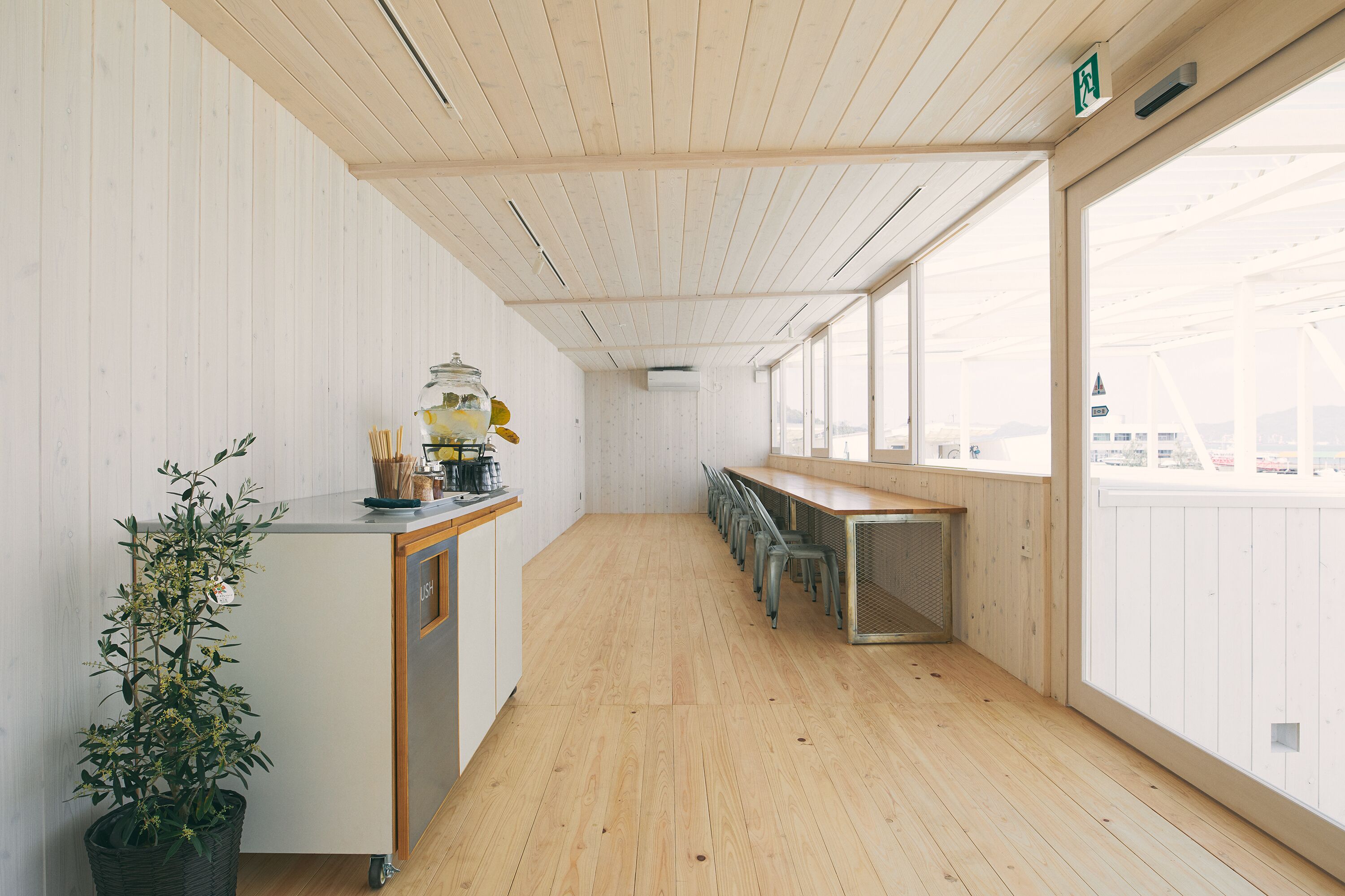 A modern, minimalist room with a wooden floor and white walls, featuring a long table and chairs, a small kitchen area with a plant, and large windows offering a view.