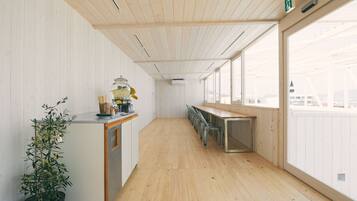 A modern, minimalist room with a wooden floor and white walls, featuring a long table and chairs, a small kitchen area with a plant, and large windows offering a view.