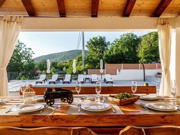 Une table à manger dressée avec des assiettes, des couverts et des verres à vin, donnant sur un espace piscine agrémenté de chaises longues et de parasols.
