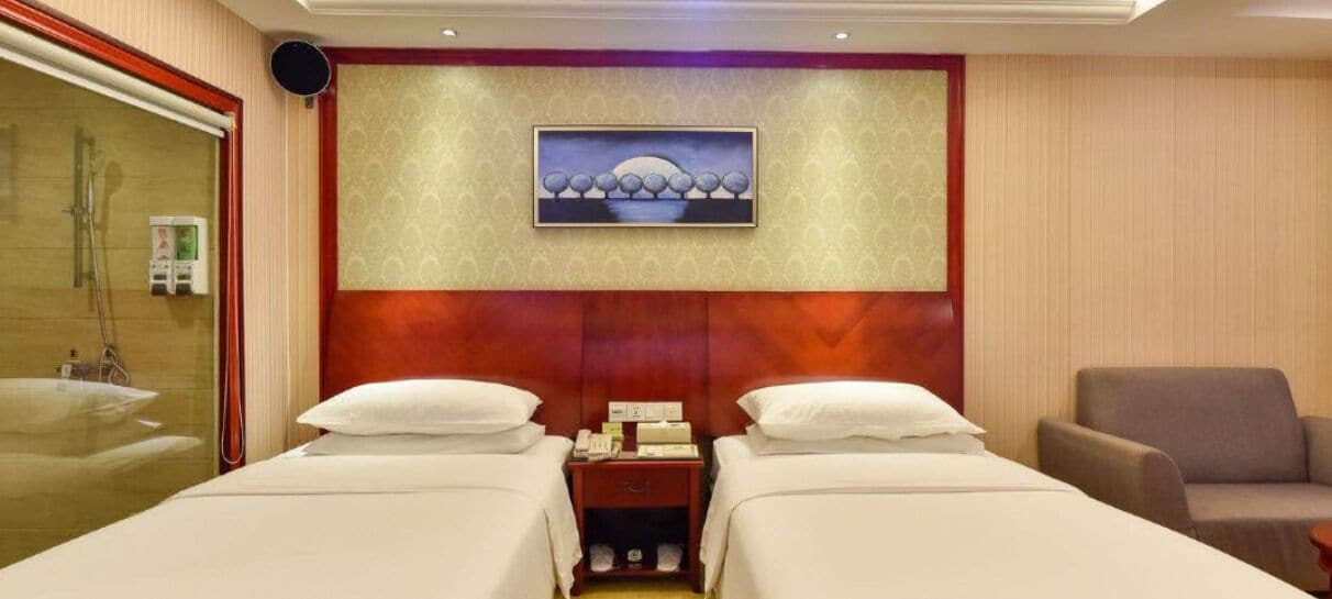 A hotel room with two beds, a wooden headboard, a nightstand, a chair, and a painting on the wall.