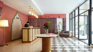 Eine Hotellobby mit einer Rezeption, einem runden Tisch mit Blumenvase, einem Schachbrettmuster Boden und großen Fenstern.