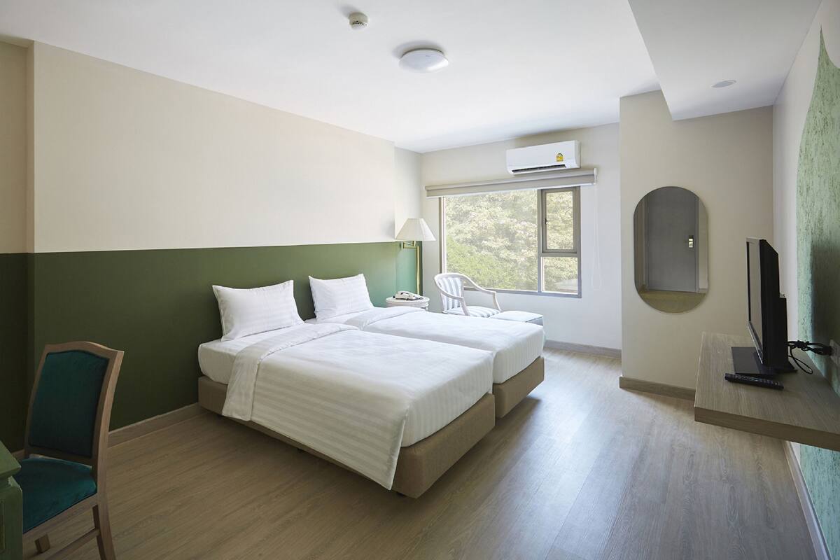 Habitació d'hotel amb dos llits, escriptori amb televisió, una cadira i finestra amb vista a arbres.