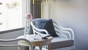 Ein weißer Stuhl mit Kissen, ein kleiner runder Tisch mit einer Vase und einem Buch sowie eine Stehlampe im Hintergrund.