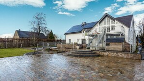 Et hus med en stenterrasse, en trappe op til en balkon og en have med hegn.