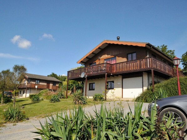 Ein zweistöckiges Holzhaus mit Balkon, umgeben von Grünpflanzen und einem parkenden Auto.