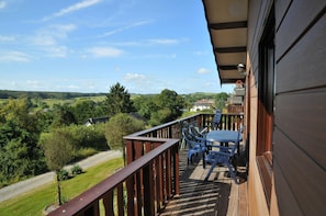 A wooden deck with a table and chairs, overlooking a green landscape with trees and a road.