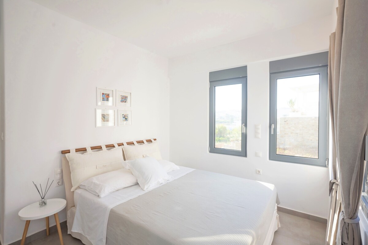 A bedroom with a large bed, a small side table, and two windows with curtains.
