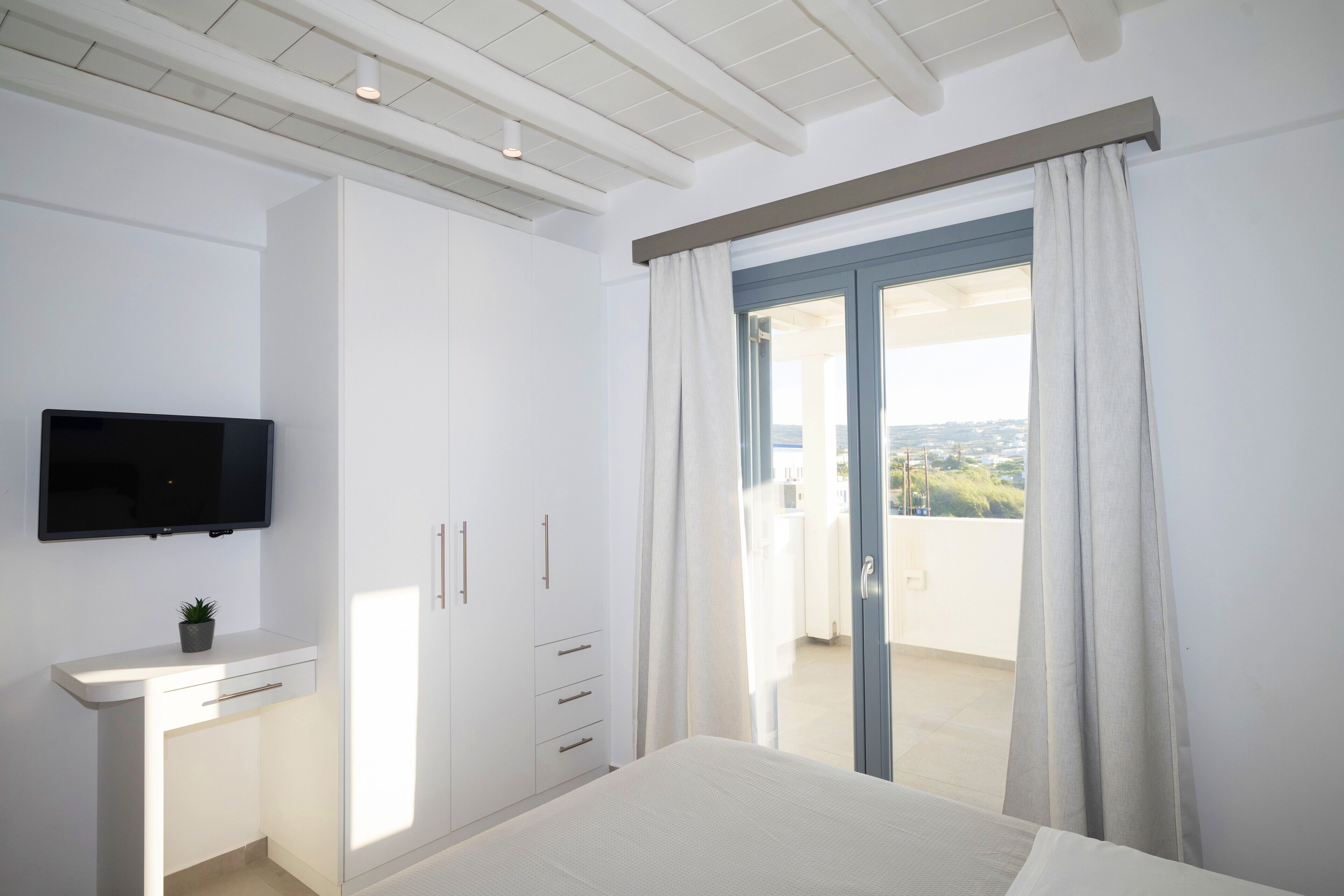 A bedroom with a bed, a TV mounted on the wall, a desk with drawers, and a view of the outdoors through a sliding door.