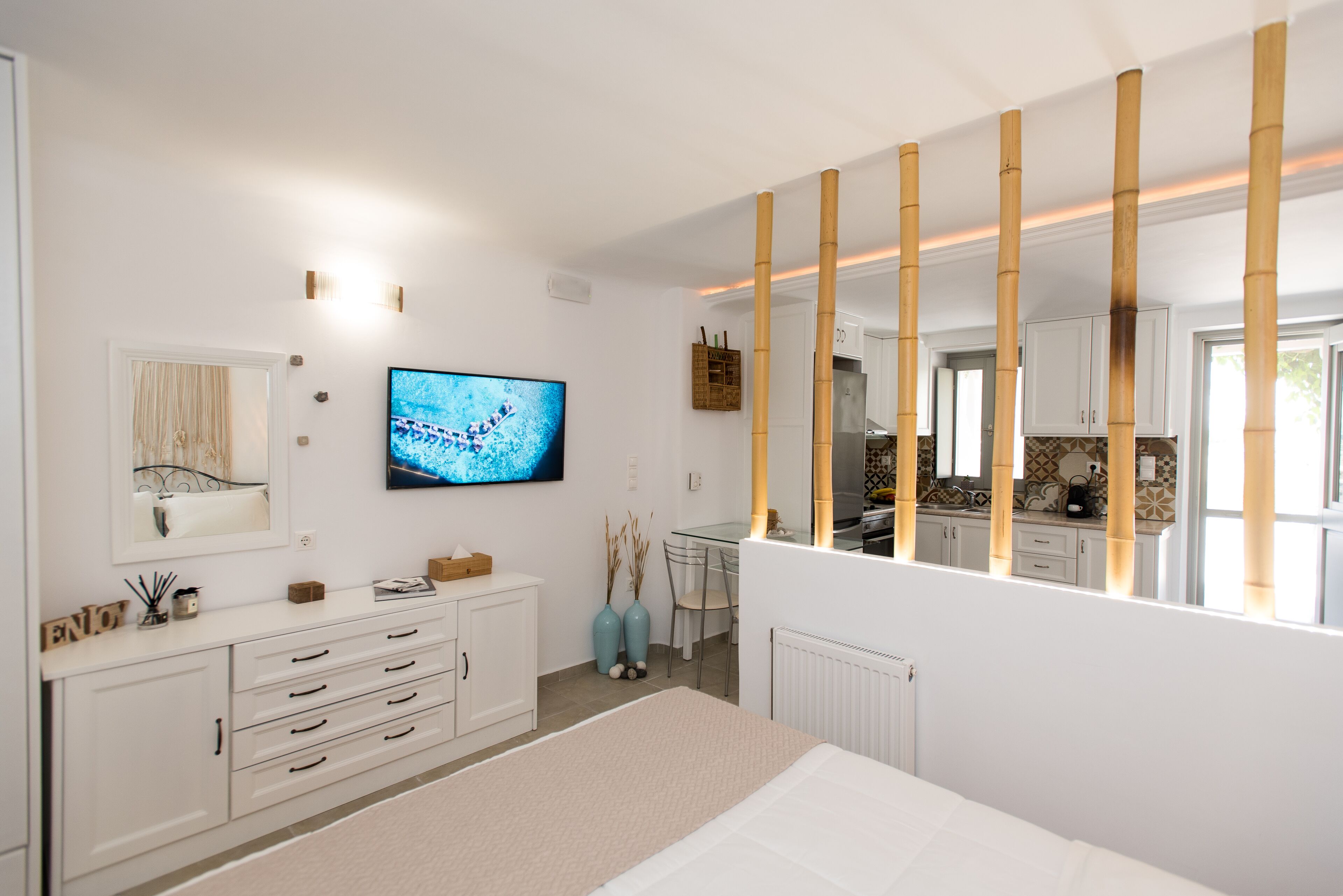 A modern bedroom with a large bed, a TV, a dresser, and a view into the kitchen.