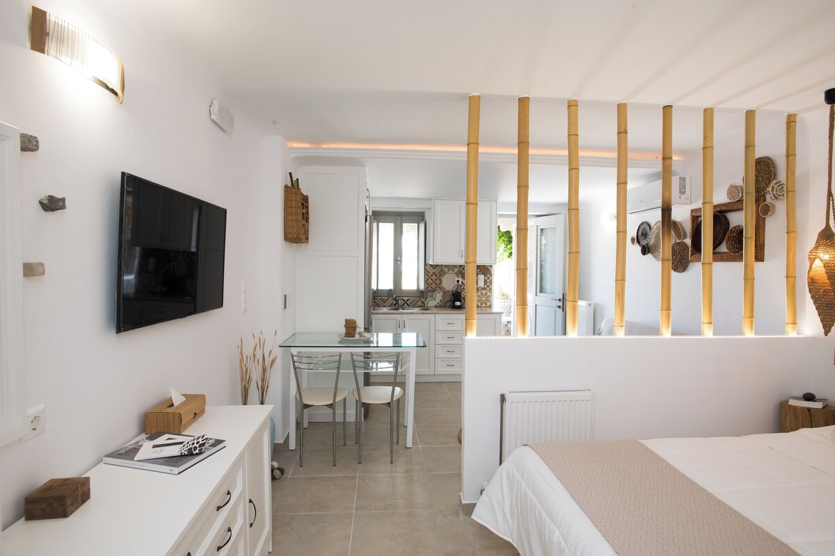A modern hotel room with a bed, a TV, a dining area with chairs, and a kitchen in the background.