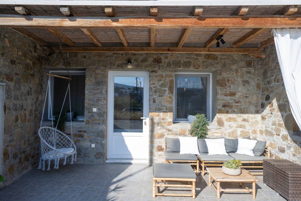 A stone building with a wooden ceiling, a white swing chair, a sofa with cushions, and a small plant on a table.
