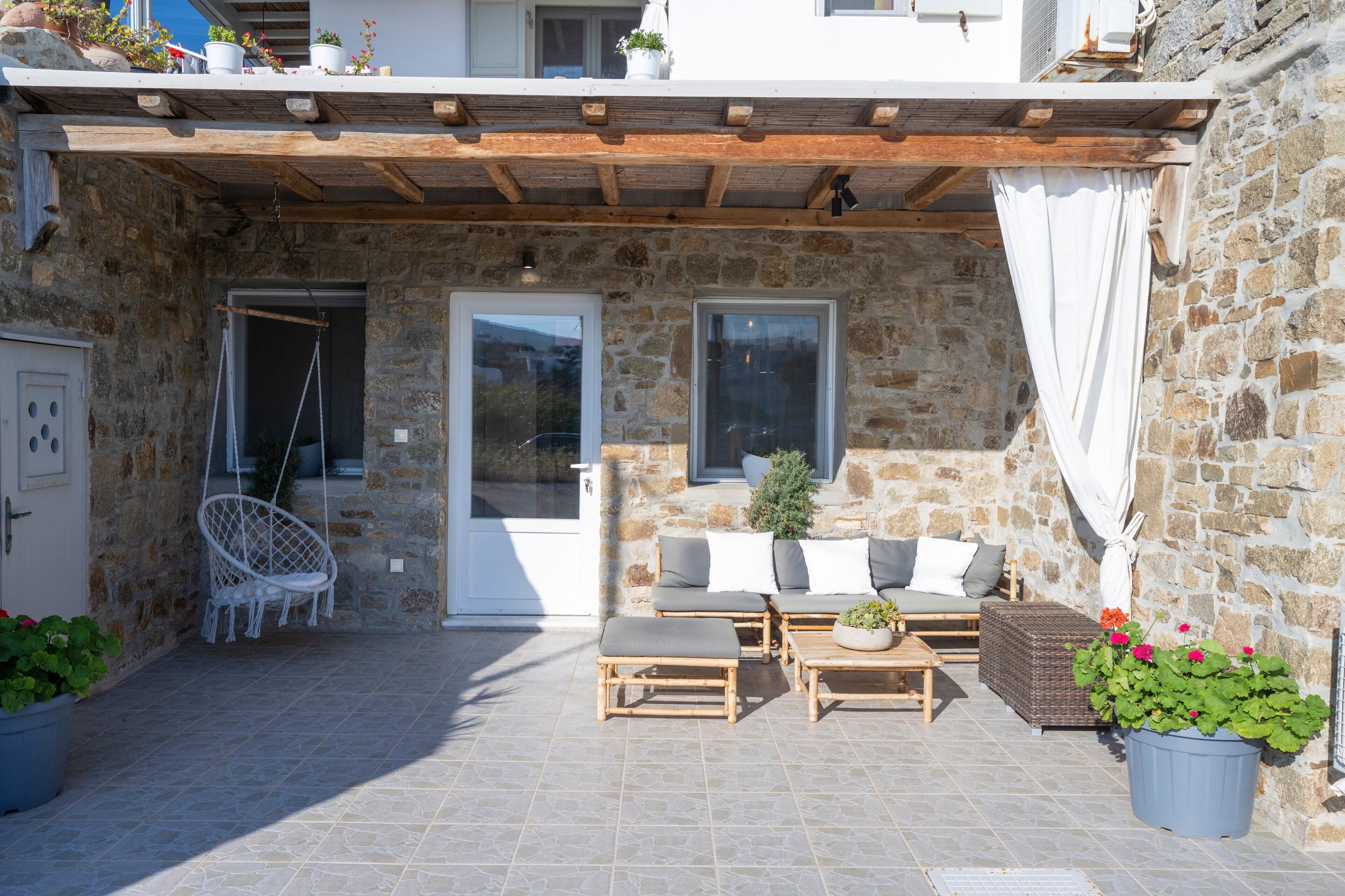 A stone building with a covered patio area featuring a sofa, two wooden chairs, a small table, and potted plants.