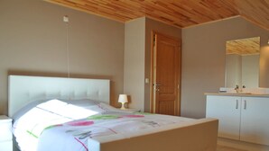 A bedroom with a bed, bedside table, lamp, wooden door, and a bathroom with a sink and mirror. - Holiday Home in Ardennes With Bubble Bath (Somme-Leuze)