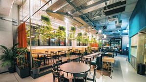 Un espace de restauration intérieur moderne, avec des tables et des chaises, des plantes en pot et une cuisine en arrière-plan.