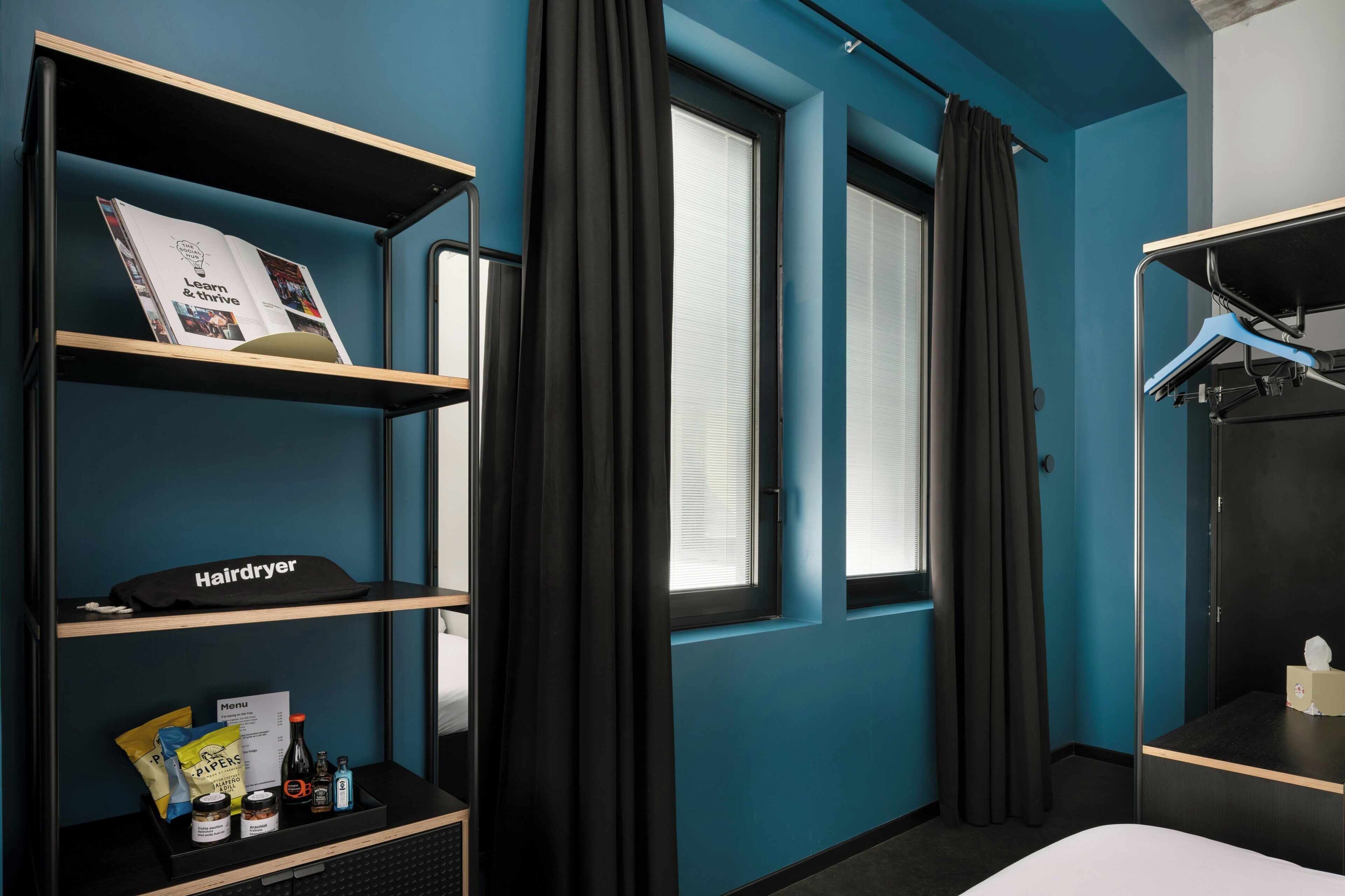 Une chambre d’hôtel avec un mur bleu, une fenêtre avec des rideaux, un lit, une étag ère avec un sèche-cheveux et une petite table avec des articles de toilette.