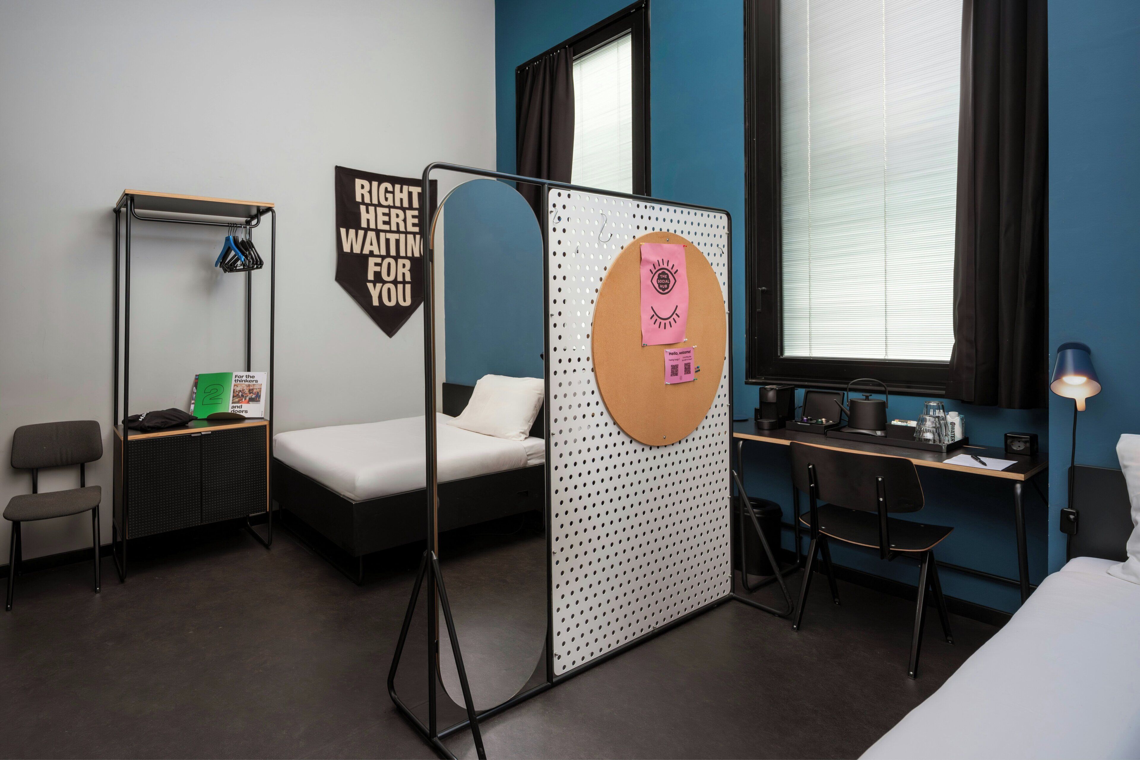 Une chambre d’hôtel équipée d’un lit, d’un bureau, d’une chaise, d’une armoire et d’un paravent orné d’affiches motivantes.