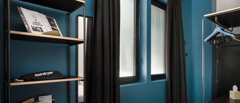 Een hotelkamer met een blauwe muur, een raam met gordijnen, een bed, een plank met een haardroger, snacks en een menukaart.